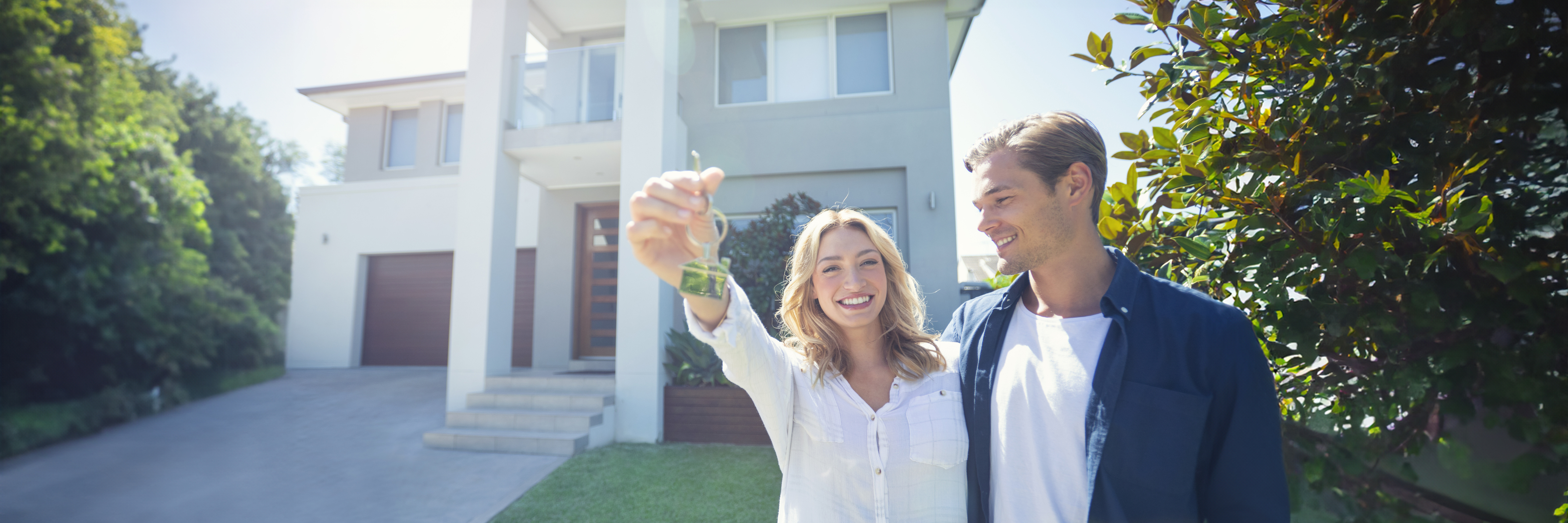 A smiling couple holding a house key in the foreground, with a modern two-story home behind them, symbolizing new home ownership.