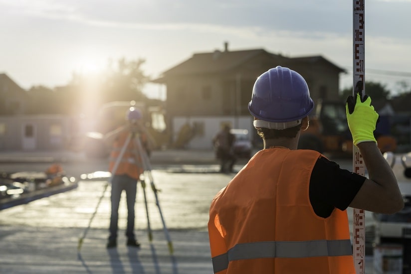 A construction worker in a reflective vest and helmet using a surveyor's level rod with another surveyor in the background using a theodolite at a construction site during sunset.