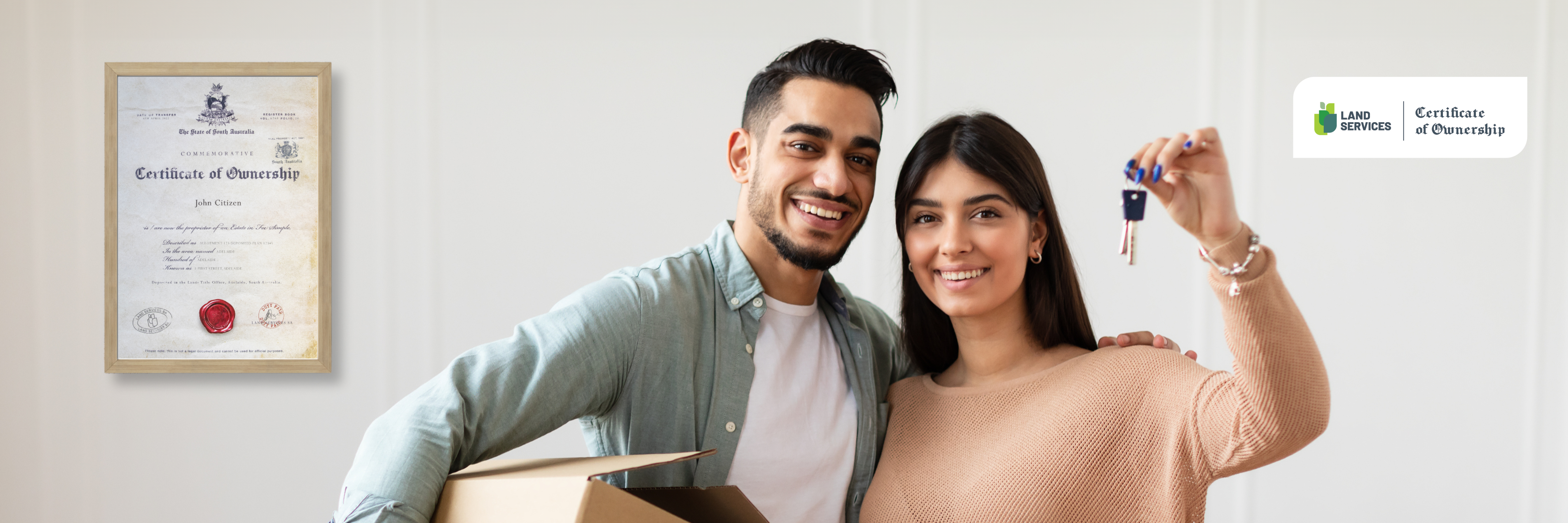 A happy young couple with a moving box, the woman holding up a set of keys, standing in front of a framed "Certificate of Ownership" on the wall, symbolizing new homeownership.